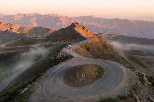 A winding dirt road switchbacking up a misty mountain pass at sunrise, with a single car climbing the ridge