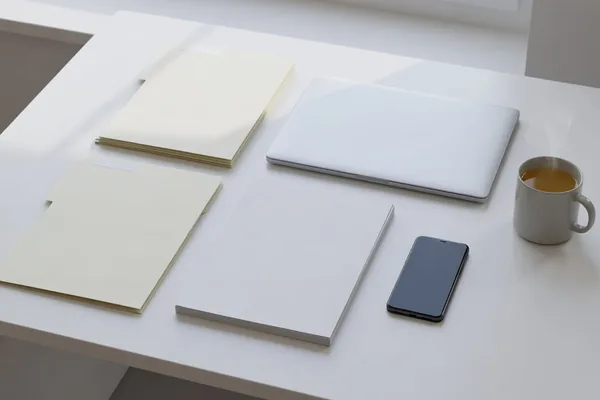 Plain folders, a blank notebook, a closed laptop, a phone and a cup of tea laid out on a bright white desk