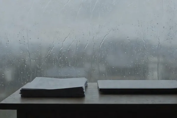 A stack of papers and a closed laptop on a table in front of a rain-streaked window
