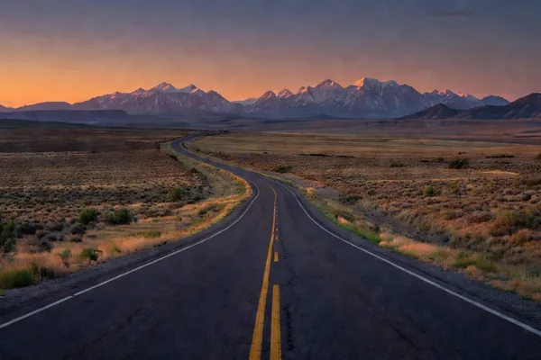 An empty two-lane highway winding across open high desert toward a snow-capped mountain range at sunset