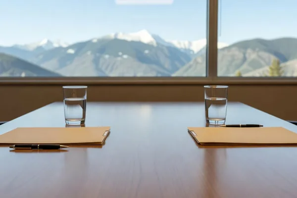 Two folders, pens and glasses of water set across a conference table with snowy mountains outside